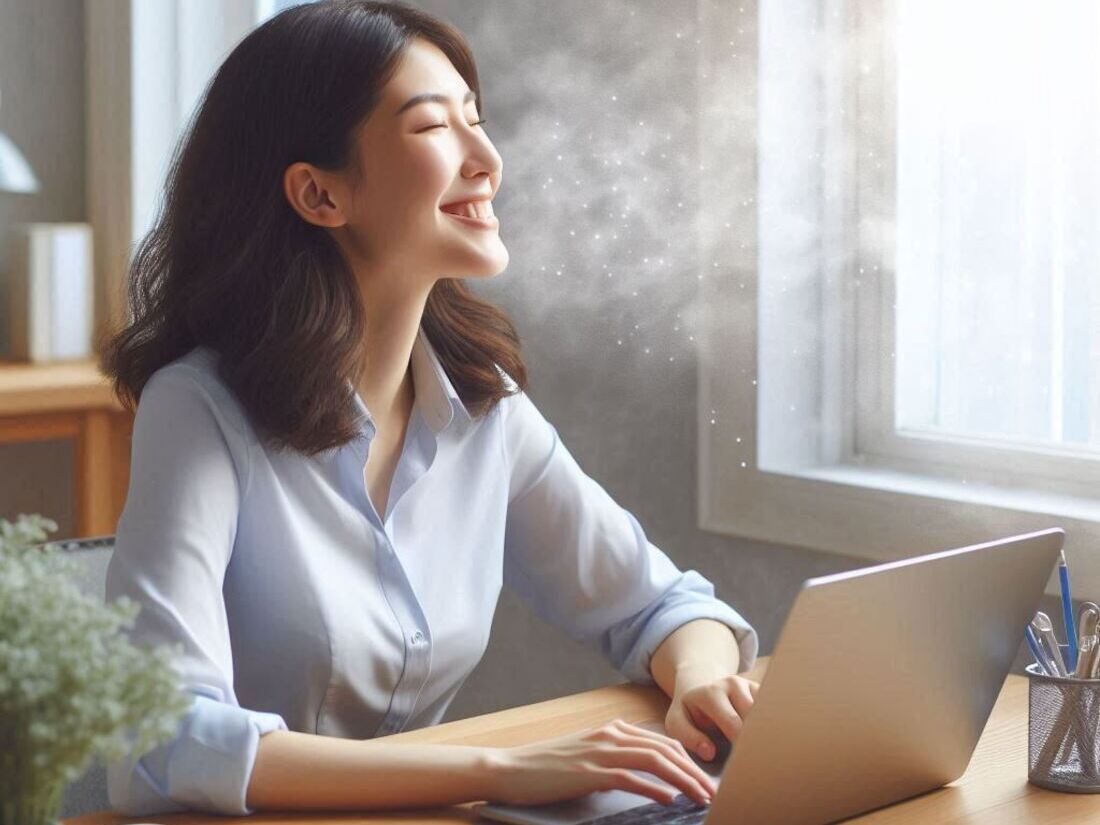 A person is seated at a desk, working contentedly on a laptop, smiling as they inhale the fresh breeze flowing in from an open window, symbolizing enhanced well-being and concentration