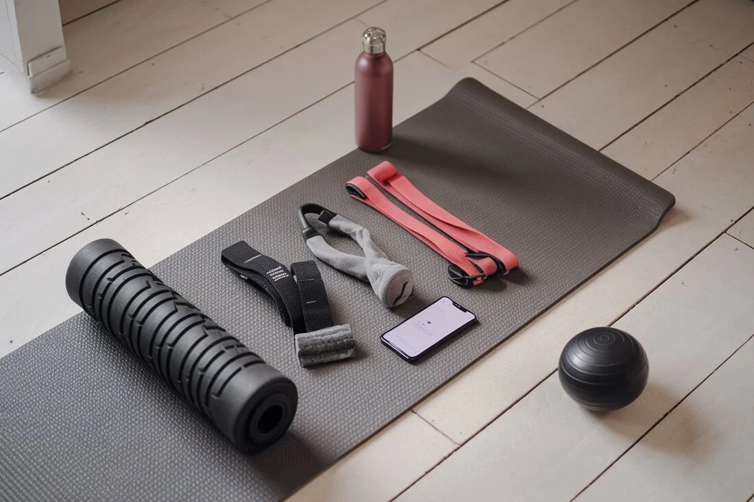 A yoga mat with neatly arranged recovery tools a foam roller, resistance bands, massage ball, water bottle, and a smartphone with a meditation app